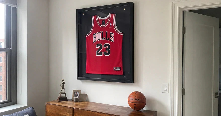 Maillot de basket rouge style Chicago Bulls numéro 23 encadré dans une vitrine murale noire, au-dessus d’un meuble en bois avec un ballon et un trophée.