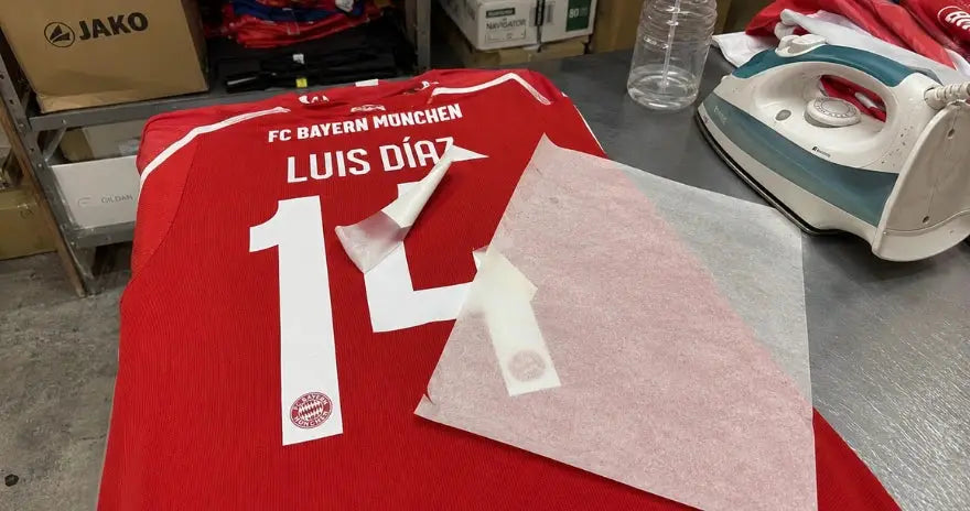 Recollage du flocage d’un maillot Bayern Munich Luis Díaz sur une table de travail, avec fer, papier de protection et spray d’eau.