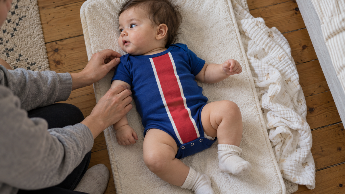 Parent ajustant les épaules d’un body de foot bleu sur son bébé pendant le change à la maison